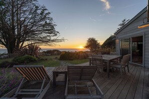 Une terrasse en bois avec une table et des chaises, d’où l’on peut admirer le coucher de soleil sur l’océan.