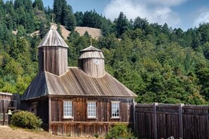 Une cabane en bois rustique, dotée d’un clocher et d’une petite construction adjacente, entourée d’une forêt dense et avec une colline en arrière-plan.