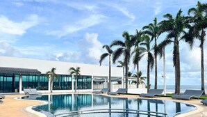 Uma área de piscina com borda azulejada, cadeiras de descanso, palmeiras e um edifício com janelas amplas.