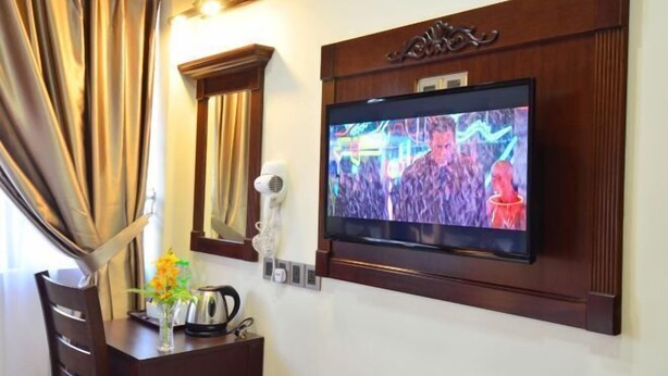 A hotel room with a flat-screen TV mounted on the wall, a hairdryer, a kettle, and a vase with flowers on a wooden table. - Hotel Kooler Inn (Kota Kinabalu)