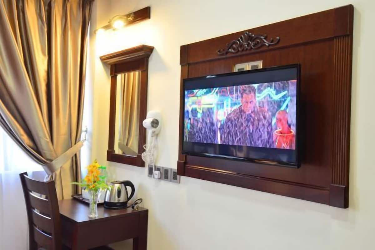 A hotel room with a flat-screen TV mounted on the wall, a hairdryer, a kettle, and a vase with flowers on a wooden table.