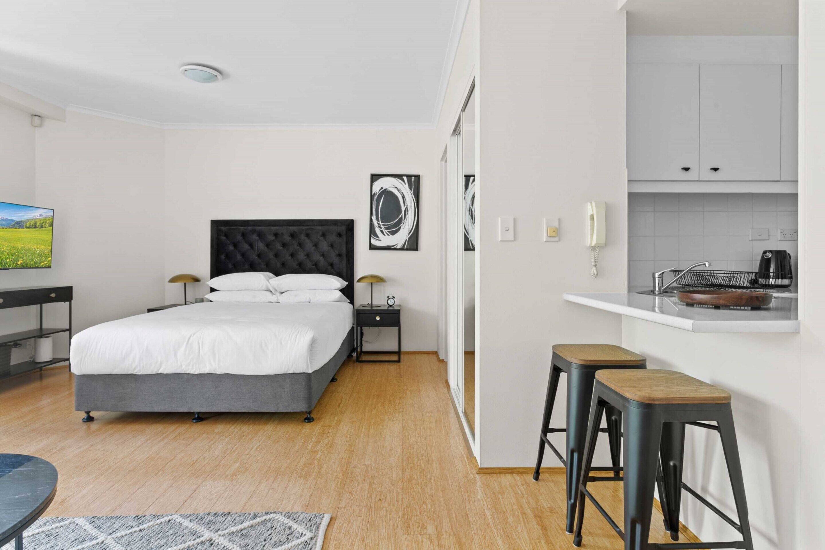 A modern hotel room with a large bed, a desk, a chair, a small kitchen area, and a view of a green landscape.