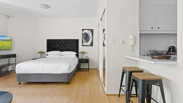 A modern hotel room with a large bed, a desk, a chair, a small kitchen area, and a view of a green landscape.