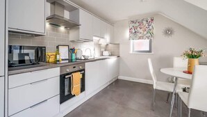 A modern kitchen with white cabinetry, a built-in oven, and a dining area with a white table and chairs. - Utility Street - 1-bed Apt + Parking (Belfast)