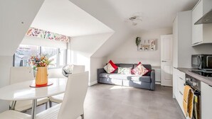 A modern kitchen with white cabinets, a microwave, and an oven. There is a dining area with a white table and chairs, and a gray sofa with colorful cushions. - Utility Street - 1-bed Apt + Parking (Belfast)