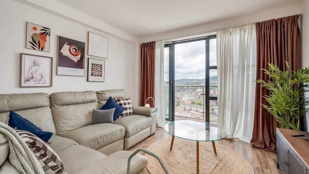 Uma sala de estar moderna com um sofá bege, uma mesa de centro de vidro e vista para um horizonte urbano através de grandes janelas.