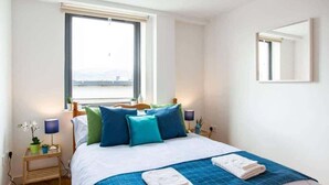 Quarto com cama, almofadas, mesa de cabeceira, abajur e janela.