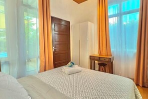 A hotel room with a bed, a wooden door, a desk with a chair, and a window with curtains.