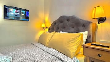 A hotel room with a tufted headboard, a flat-screen TV mounted on the wall, a bedside lamp, and a neatly made bed with patterned bedding.