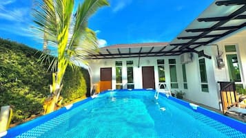 A swimming pool with a clear blue water surface, surrounded by a white building with large windows and a covered patio area.