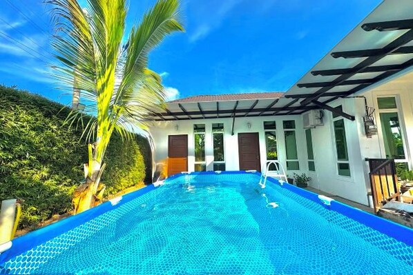A swimming pool with a clear blue water surface, surrounded by a white building with large windows and a covered patio area.