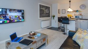 A modern living room with a flat-screen TV, a laptop, a coffee table, and a sofa. There is a kitchen area with a microwave, a sink, and a clock on the wall. - Deluxe Apartment at Central Belfast Apartments (Belfast)