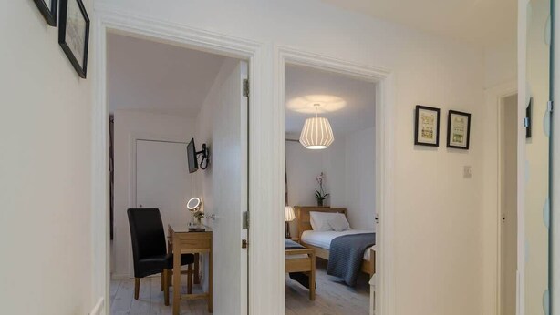 A room with a bed, bedside table, lamp, and a chair. There is a door leading to another room with a table and chairs. - Deluxe Apartment at Central Belfast Apartments (Belfast)