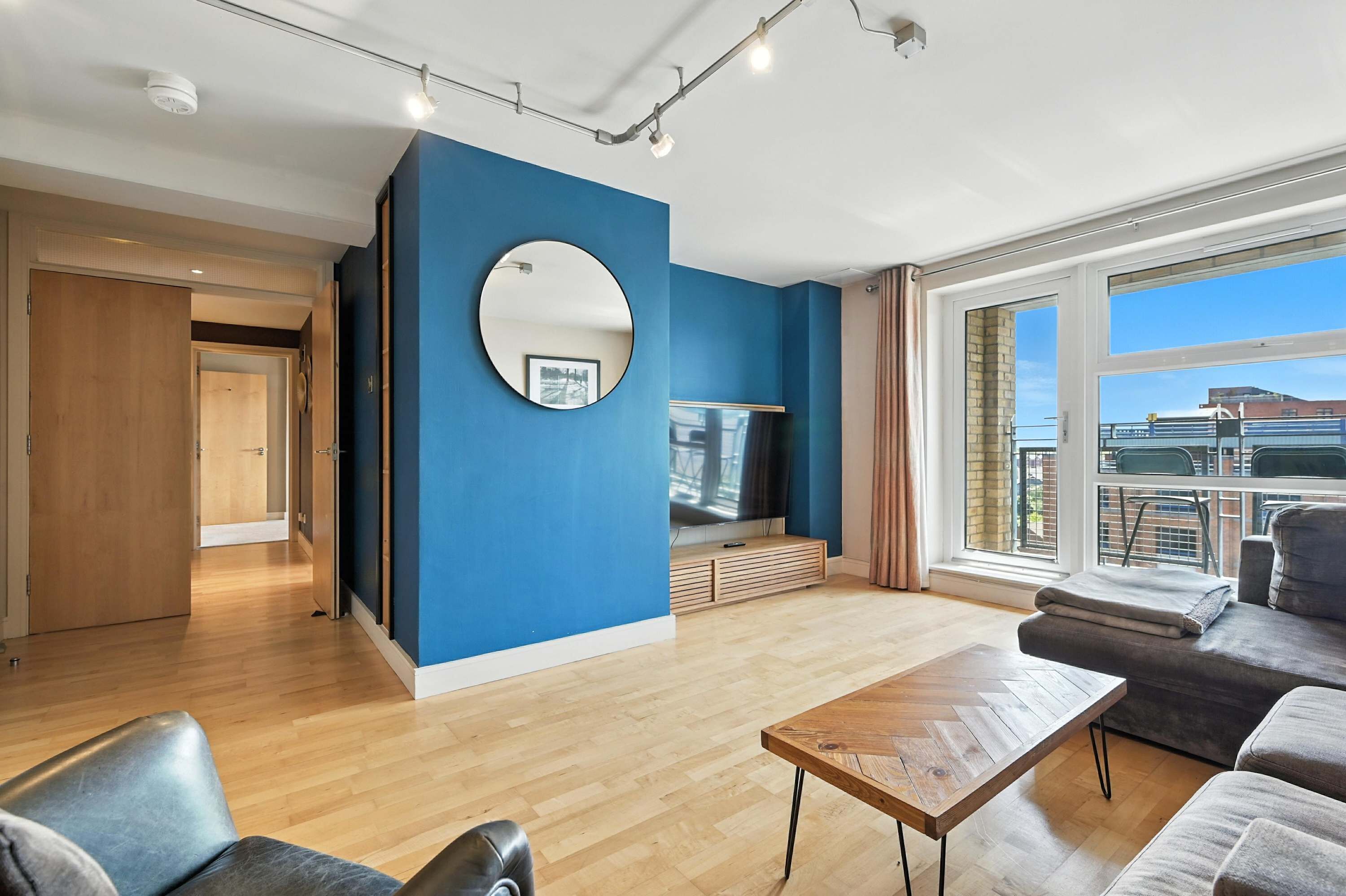 A modern living room with a blue accent wall, a round mirror, a flat-screen TV, a wooden coffee table, and a sofa.