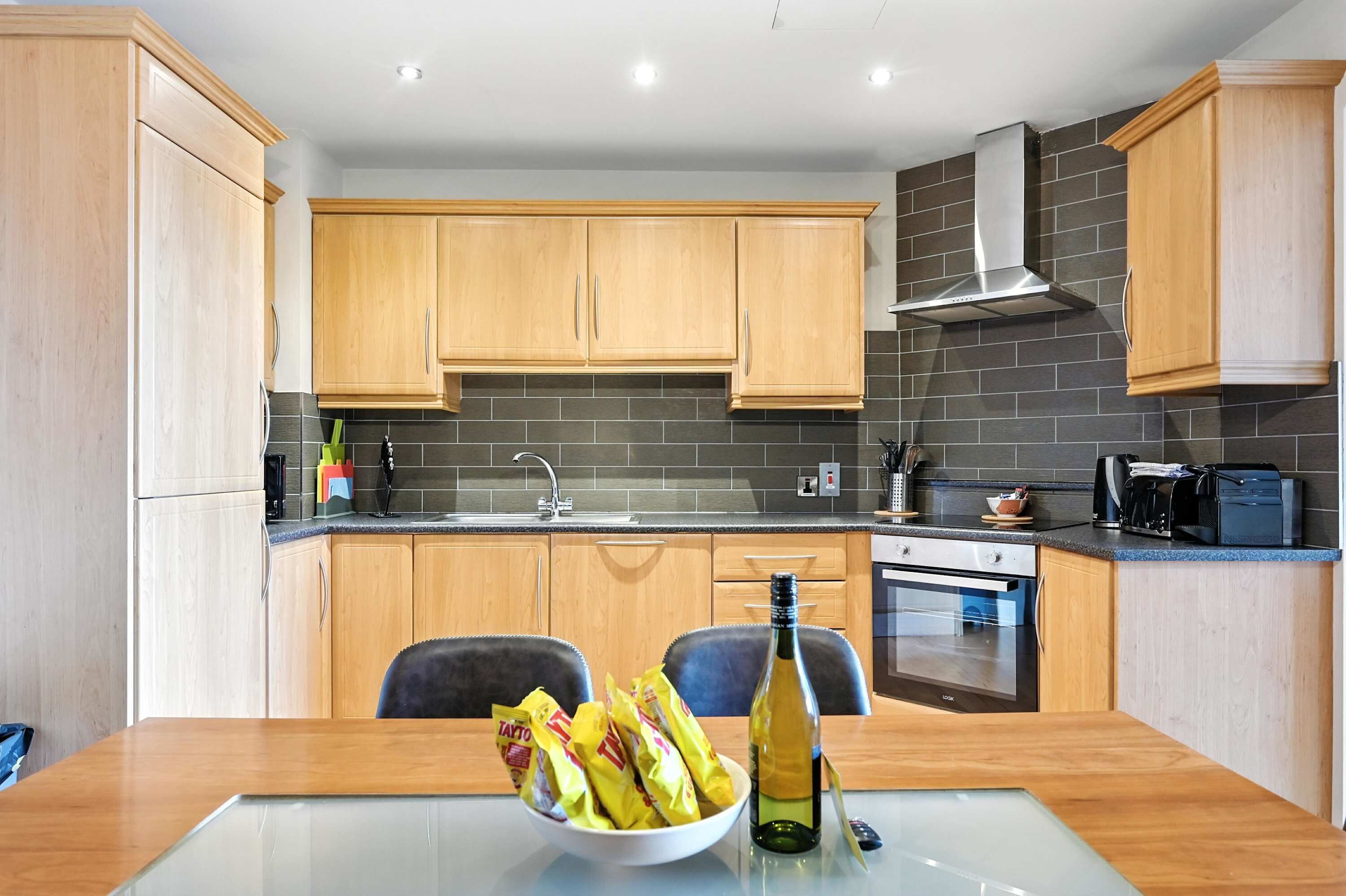 A modern kitchen with wooden cabinets, a black tiled backsplash, a stainless steel range hood, and a dining area with a table and chairs.