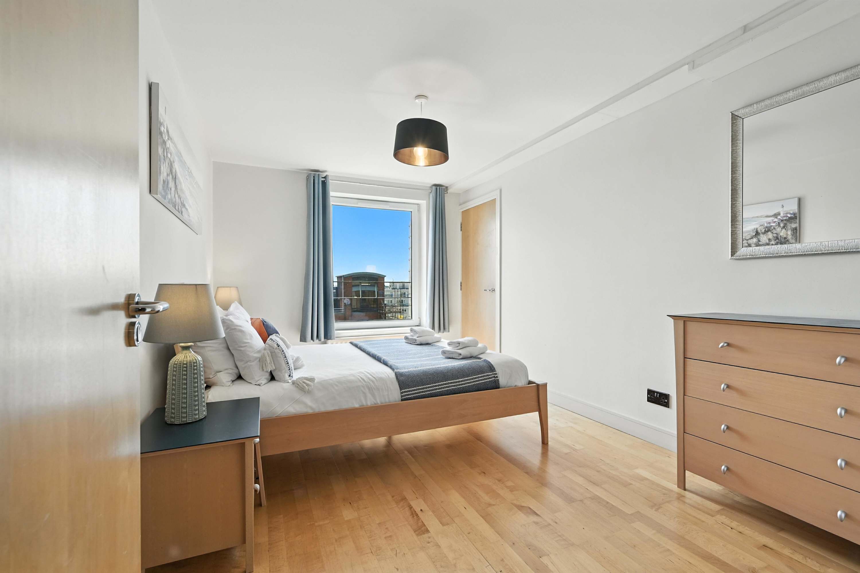 A modern bedroom with a wooden bed, a nightstand, a dresser, and a large window with curtains.