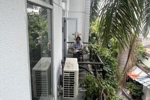 A balcony with a person seated at a table, surrounded by greenery and a building facade. - Kim’s House Entire Villa 6-BR Near My Khe Beach (Da Nang)