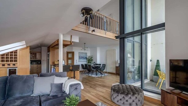 A spacious living area with a sofa, ottoman, and a coffee table. A balcony with a railing and a television is visible. - 5-bedroom apartment with river views - Goliath (Belfast)