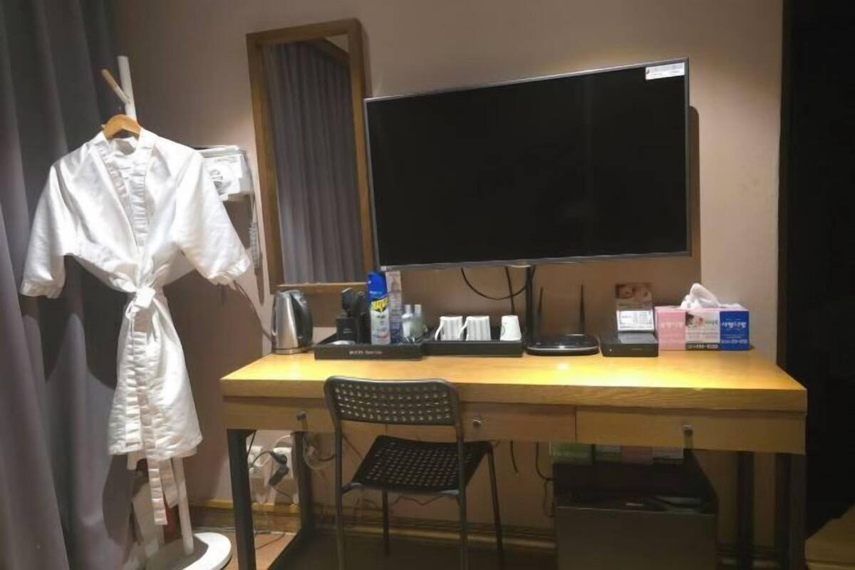 A hotel room with a wooden desk, a black chair, a white robe hanging, a flat-screen TV, and a mirror.