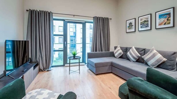 A modern living room with a grey sectional sofa, a small side table, and a television on a stand. There are three framed pictures on the wall and a window with curtains.