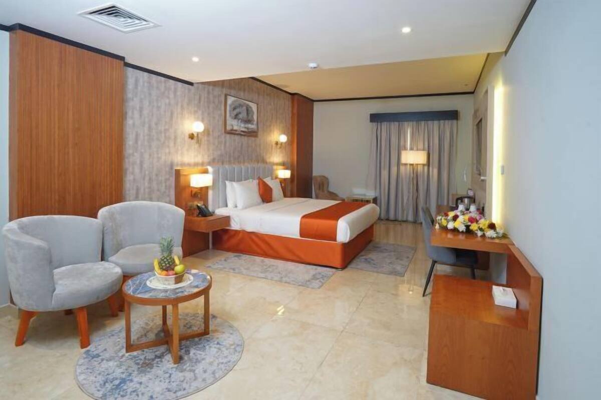A hotel room with a bed, two armchairs, a small table with fruit, a desk with a lamp, and a mirror.
