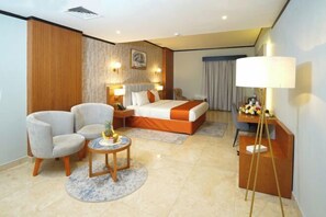 A hotel room with a bed, two armchairs, a small table with a fruit bowl, a desk with a lamp, and a wall-mounted TV. - Concorde Palace Hotel (Dubai)