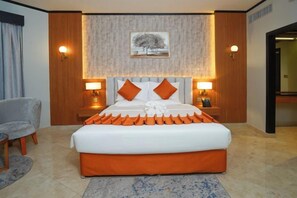 A hotel room with a large bed, two bedside lamps, a headboard, a nightstand, a chair, and a painting on the wall. - Concorde Palace Hotel (Dubai)