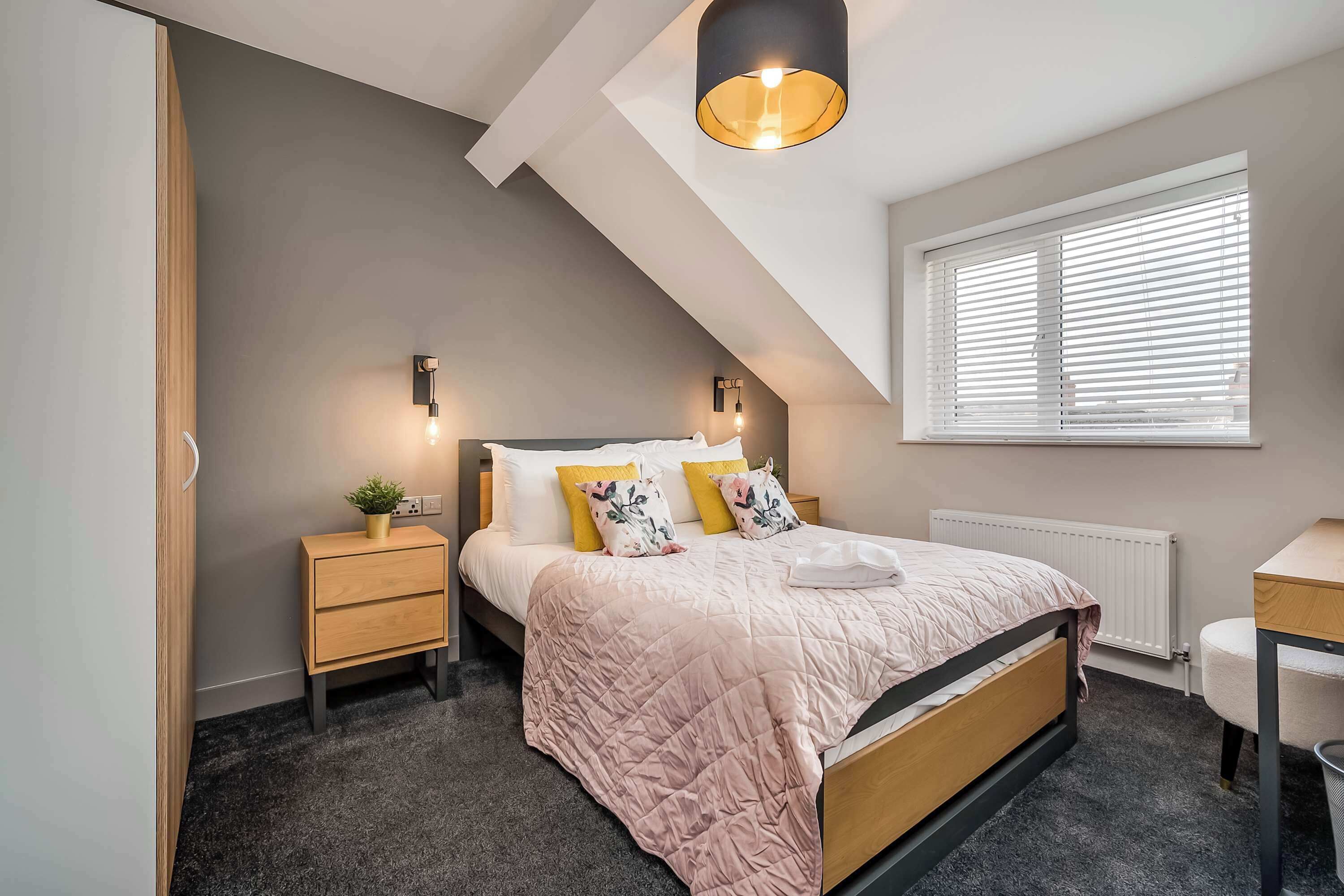 A bedroom with a sloped ceiling, a bed with a pink quilt, bedside tables, a small plant, and a window with blinds.