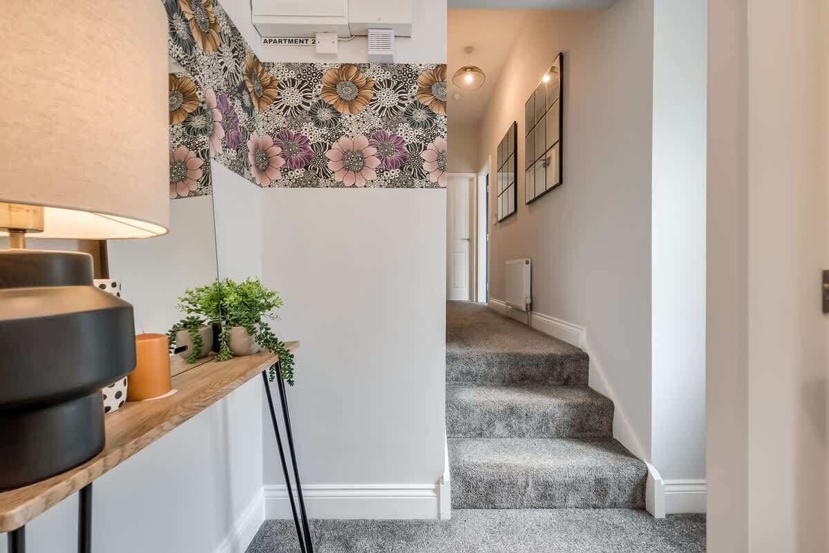 Un escalier orné d’un papier peint à motifs floraux, une console en bois avec une lampe et une plante en pot.