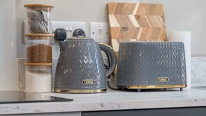 A kitchen counter with two gray kettles, a coffee maker, a jar of sugar, and a wooden cutting board.