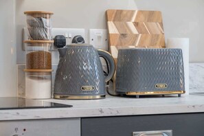 Una encimera de cocina con dos hervidores grises, una cafetera, un tarro de azúcar y una tabla de cortar de madera.