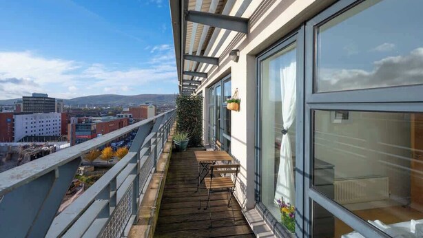 Un balcone con pavimento in legno, un tavolo e sedie, con vista su un panorama urbano che include edifici e un cielo sereno.