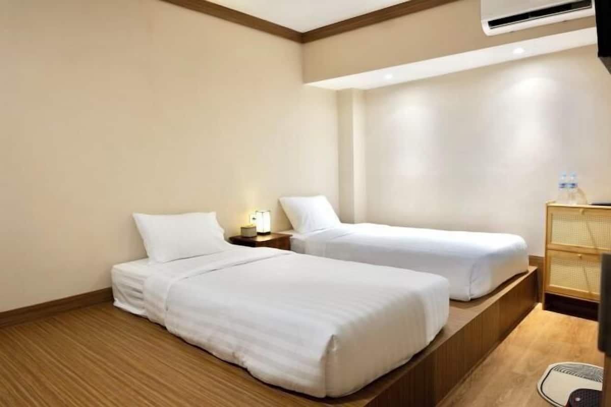 Quarto de hotel com duas camas de solteiro, uma mesa de cabeceira com abajur, um armário de madeira pequeno e uma janela com vista para o exterior.