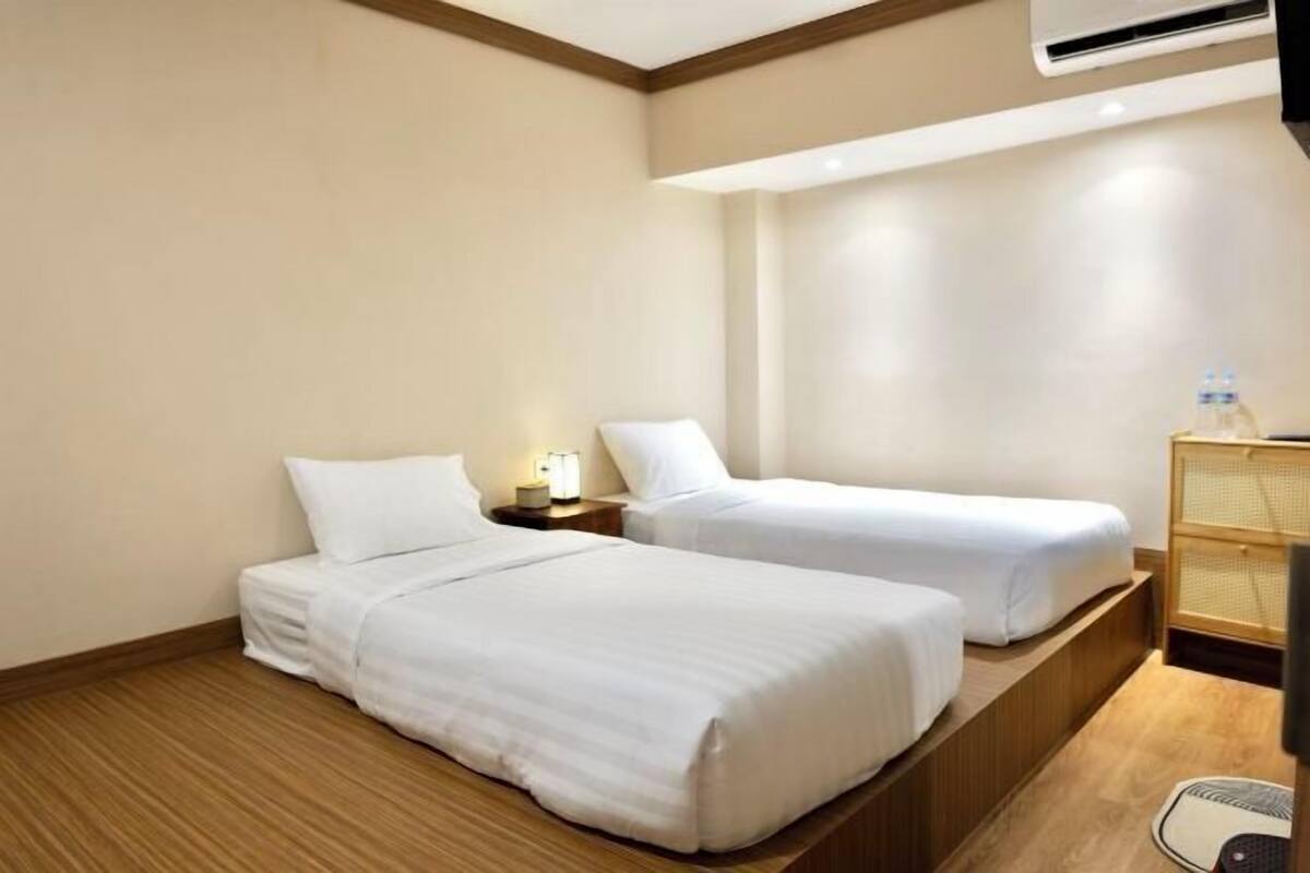 A hotel room with two single beds, a bedside table with a lamp, a small wooden cabinet, and a window with a view of the outside.
