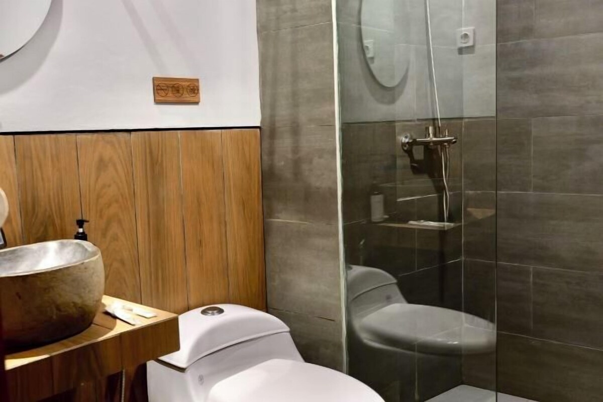 A modern bathroom with a wooden vanity, a large stone sink, a toilet, and a glass-enclosed shower area.