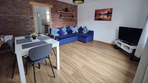 A modern living room with a brick wall, a blue sofa, a dining table with chairs, a TV stand, and a hanging light fixture.