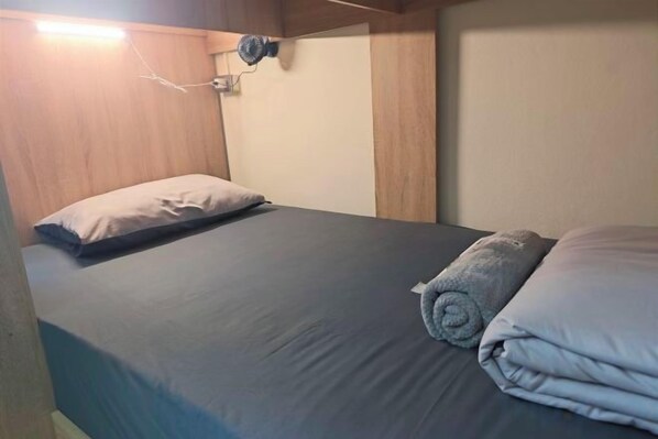 A single bed with a grey mattress and pillows, a bedside table with a lamp, and a wall-mounted fan. - Panda Lodge Home Stay (Bangkok)