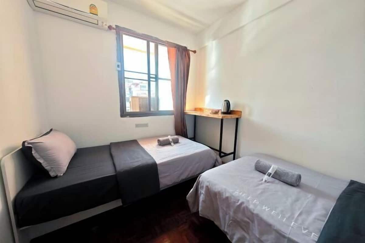 A small room with two beds, a window with curtains, a table with a kettle, and an air conditioning unit.