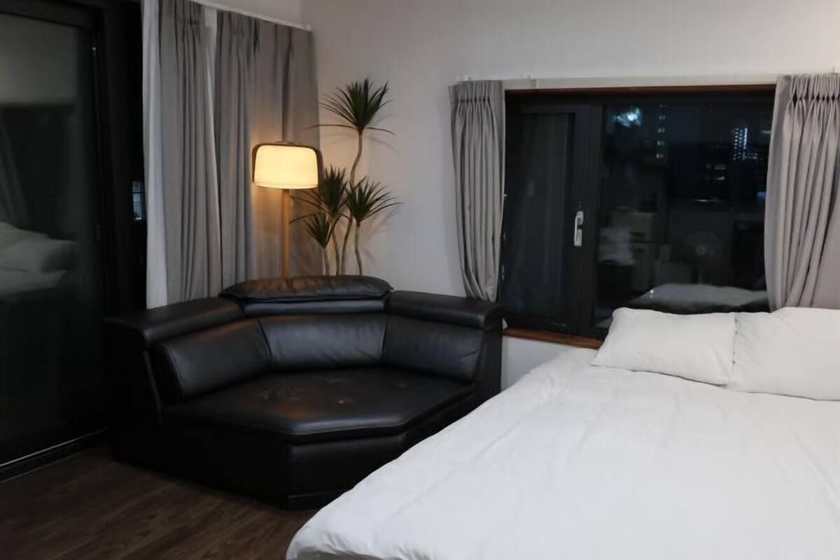 A modern hotel room with a large bed, a black leather sofa, a floor lamp, and a view of the city at night.