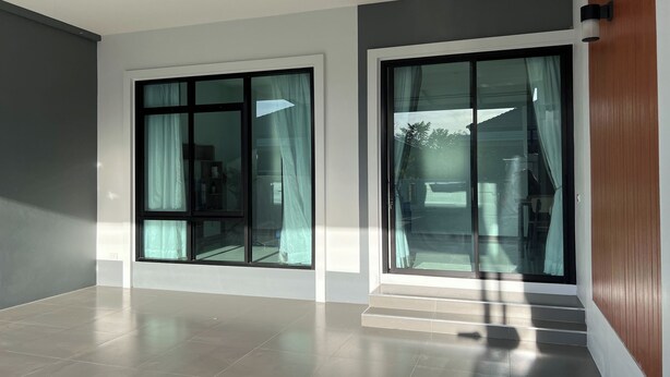 A spacious room with large sliding glass doors, a tiled floor, and a wooden panel on the right wall. - Poome House By Zuzu (Hat Yai)