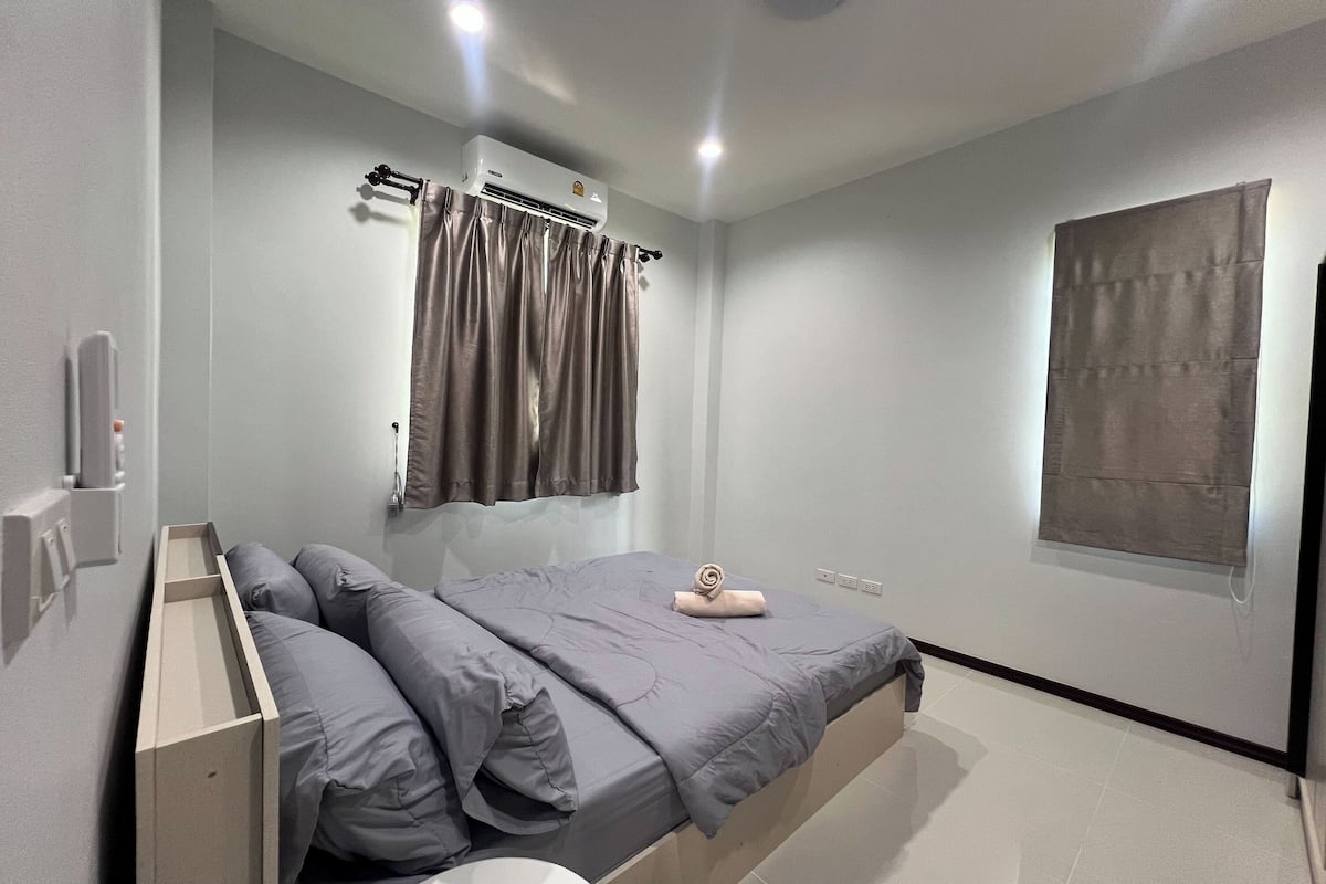 A modern bedroom with a bed, a built-in bedside table, a wall-mounted air conditioner, and a window with curtains.