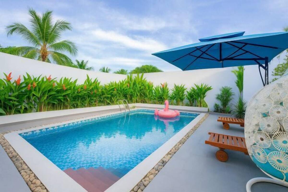 A swimming pool with a pink flamingo float, surrounded by palm trees and a blue umbrella.