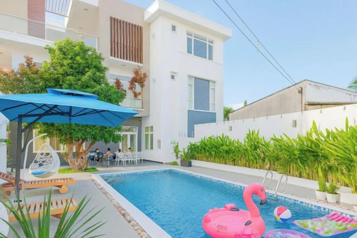 Una piscina con un flotador de flamenco rosa, una sombrilla azul y una zona de estar con una silla y una mesa.