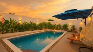 Une piscine avec un parasol bleu, une chaise longue et une petite table sur laquelle est posée une bougie.