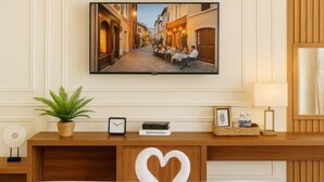 Une chambre d’hôtel avec un lit, un téléviseur fixé au mur, une table de chevet avec une plante, une horloge et une décoration en forme de cœur sur le lit.