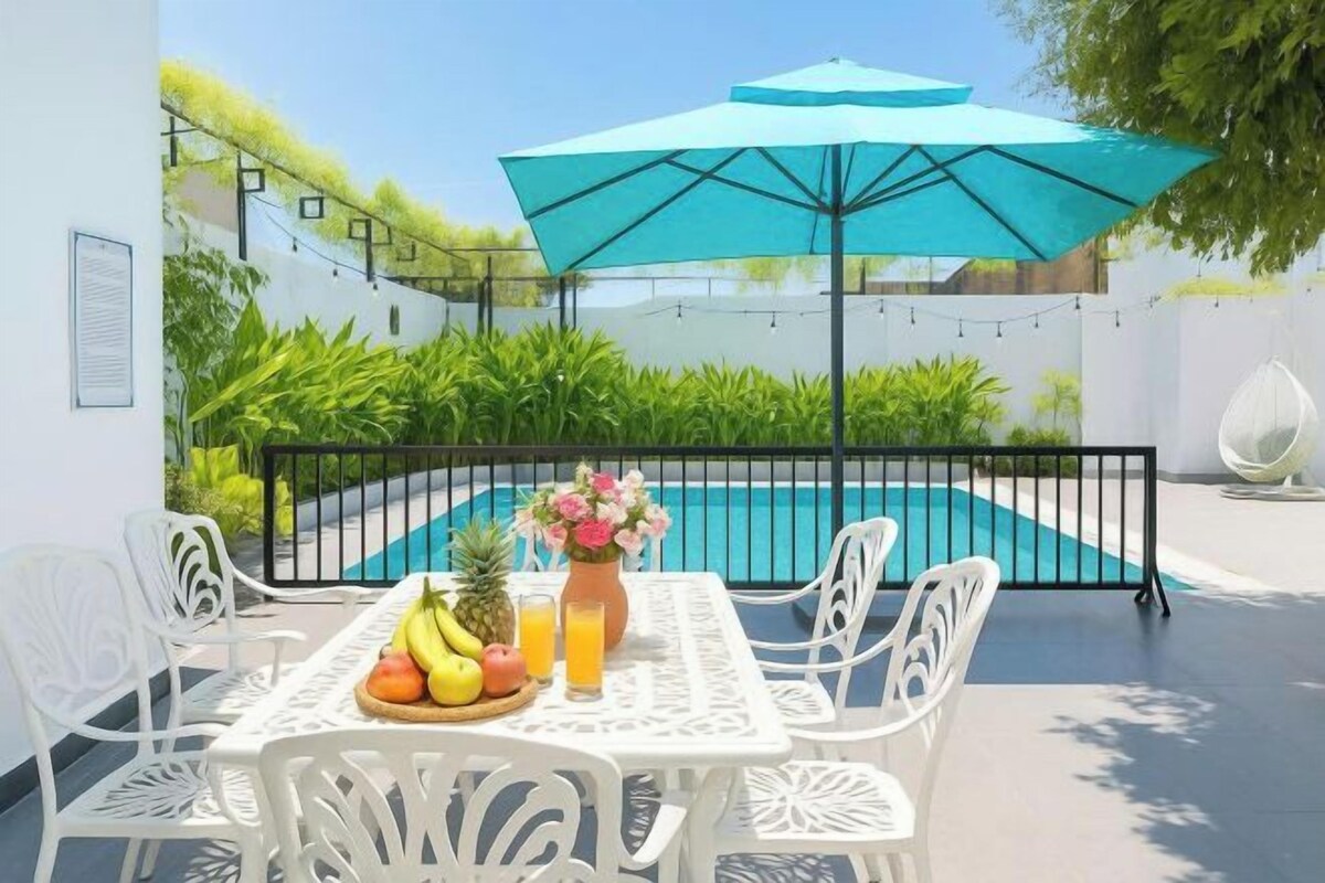 Mesa junto a la piscina con frutero, jarra y botella, rodeada de sillas blancas.