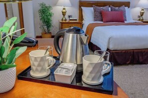 Habitación de hotel con cama, mesitas de noche, bandeja de café con tazas y tetera, y una planta en maceta.