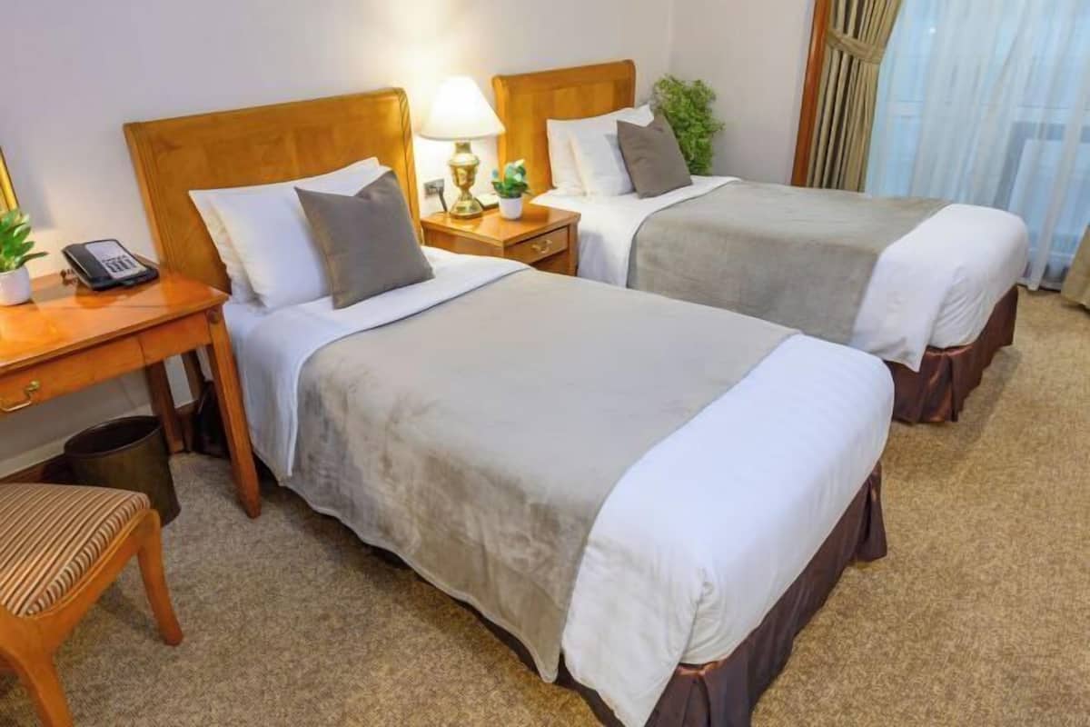 A hotel room with two beds, a desk with a lamp, a telephone, and a small chair.