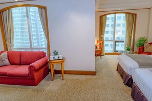 Habitación de hotel con sofá rojo, una mesita redonda, cama y un ventanal con cortinas.