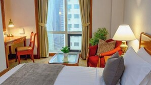 Un cuarto de hotel con cama, escritorio, silla, una mesita con una planta y vista a edificios a través de la ventana.